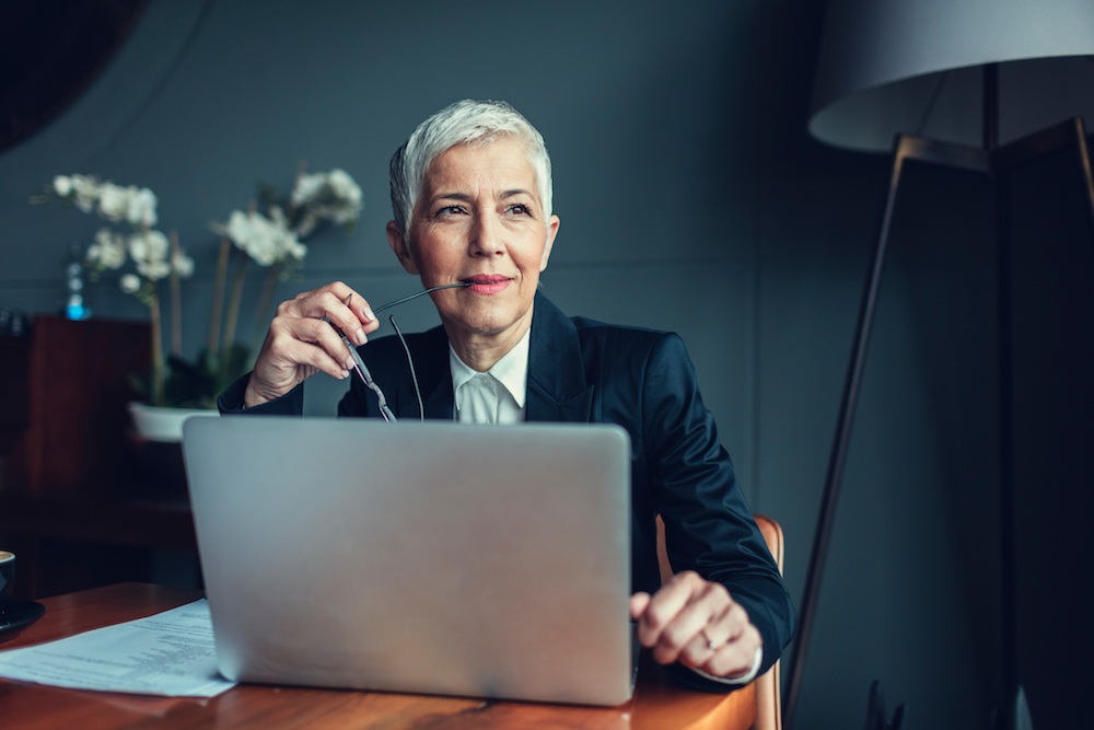 Portrait of mature businesswoman, sitting in her office and thinking. Holding eyeglasses in her mouth. In front of her on the desk is laptop.