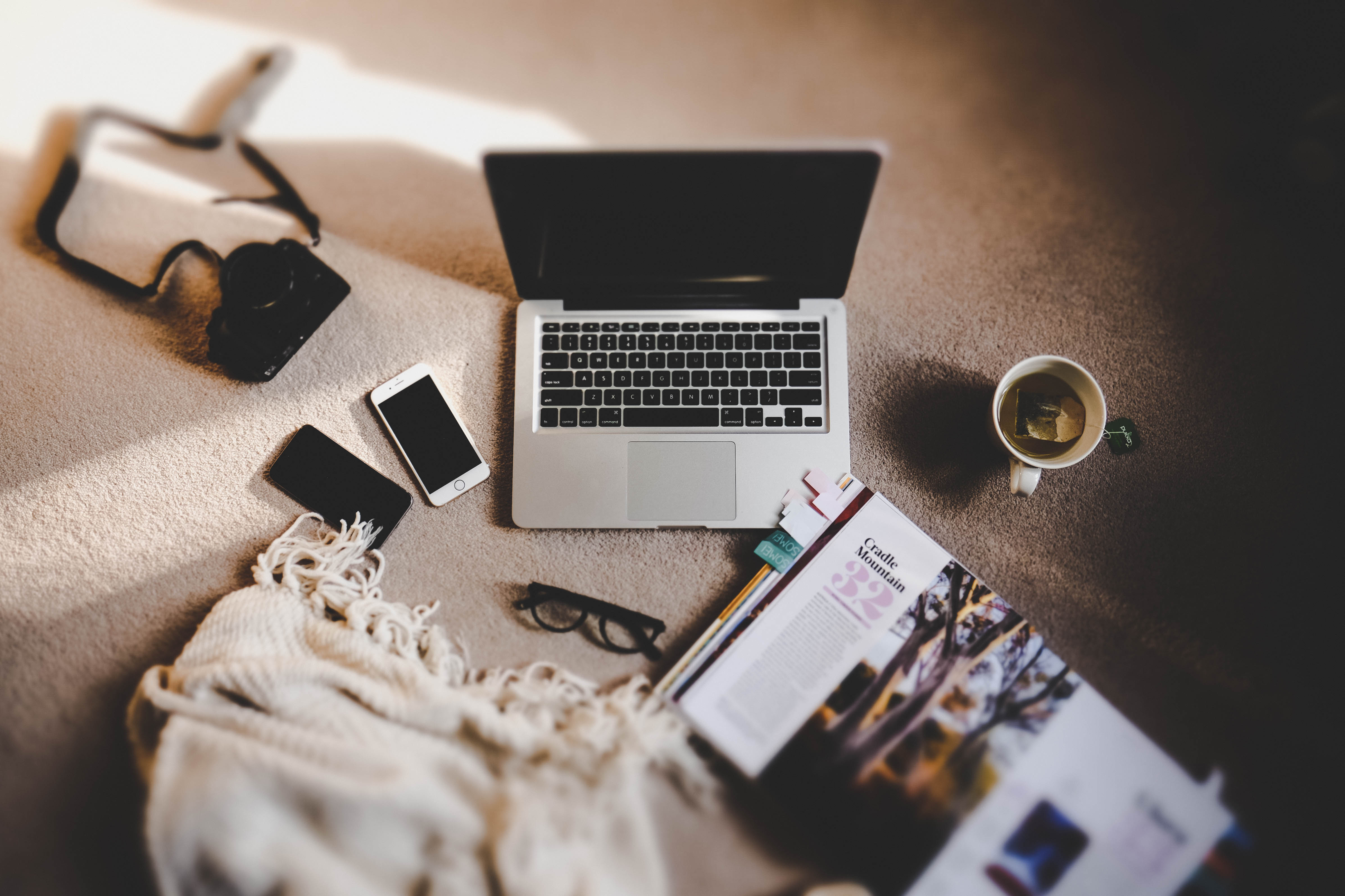 A laptop, smarthphone, camera, coffee and open magazine on the floor.