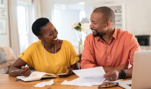 Cheerful mature couple sitting and managing expenses at home. Happy african man and woman paying bills together and managing budget. Black smiling couple checking accountancy and bills while looking at each other. (Cheerful mature couple sitting and m