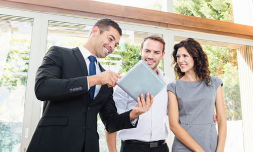 A real estate agent shows two clients listing information on a tablet.